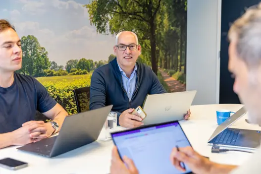 IT-expert aan tafel met twee collega’s in bespreking, kijkt vriendelijk in de camera tijdens overleg over toekomstige mogelijkheden.