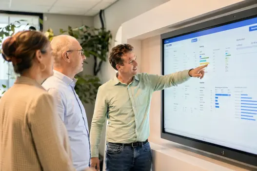 Een moderne, lichte kantooromgeving waarin medewerkers van De Alliantie samenwerken rondom een scherm met plannings- en kwaliteitsinformatie. De sfeer is professioneel, positief en mensgericht. Geen geposeerde stockfoto, maar een authentiek beeld dat samenwerking, eigenaarschap en klantgerichtheid uitstraalt.